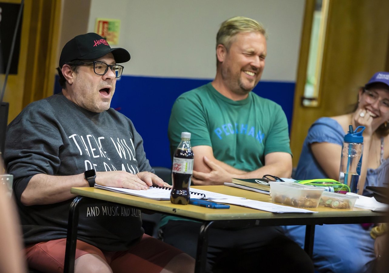 Andy Fickman and Gary Lloyd sit behind a desk both reacting to something they are seeing with an open mouth and a smile