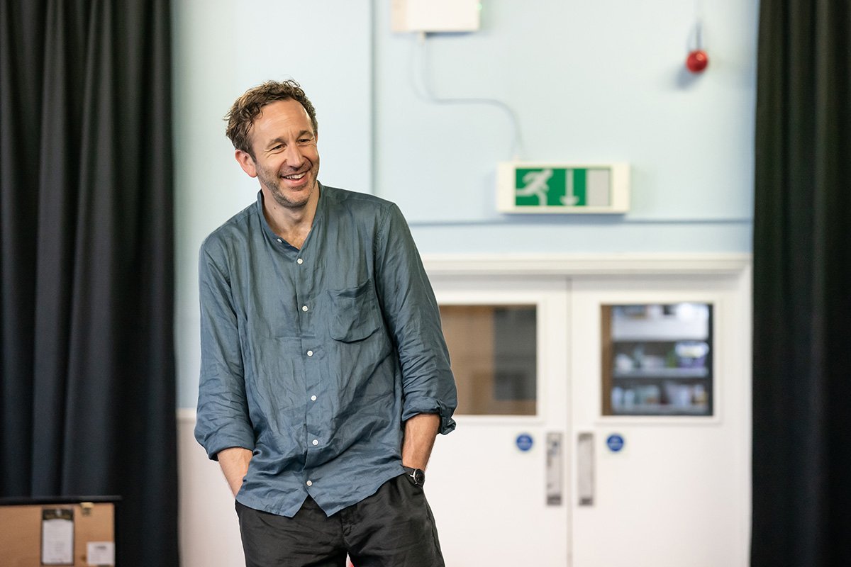 Chris O'Dowd stands with his hands in his pockets. He is smiling (almost laughing).