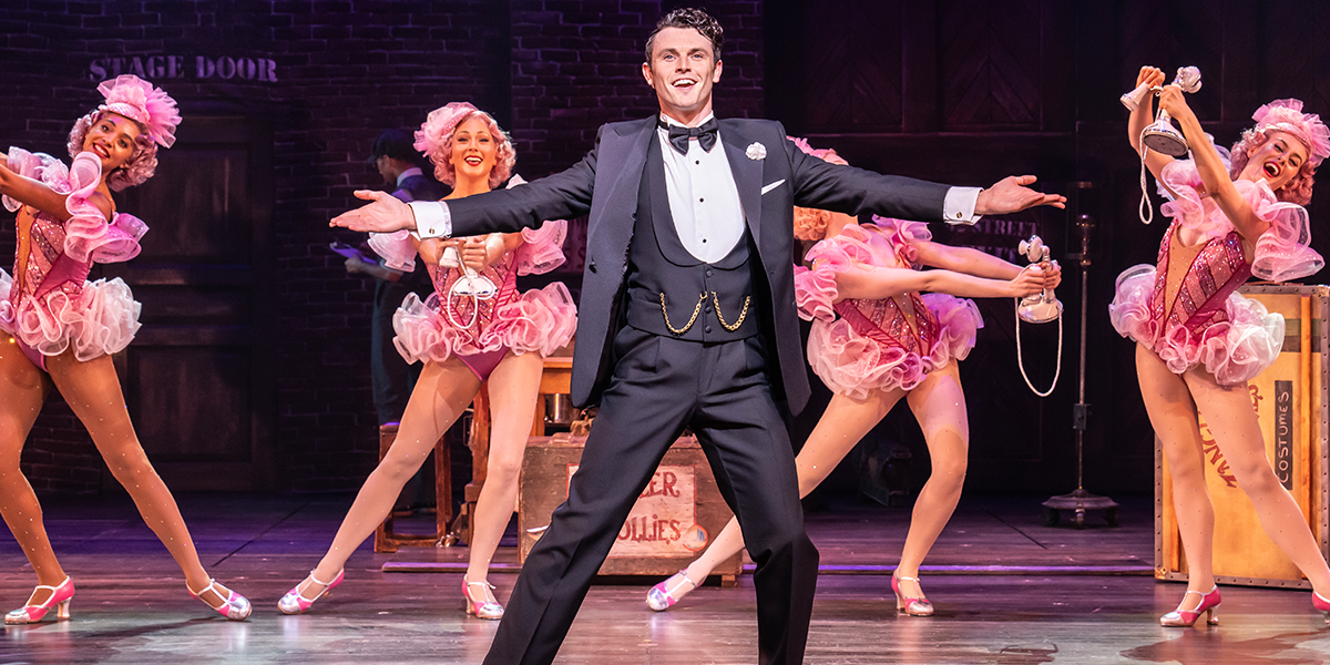 Charlie Stemp as Bobby Childs in a suit, while the Zangler Follies dance behind him in pink outfits.