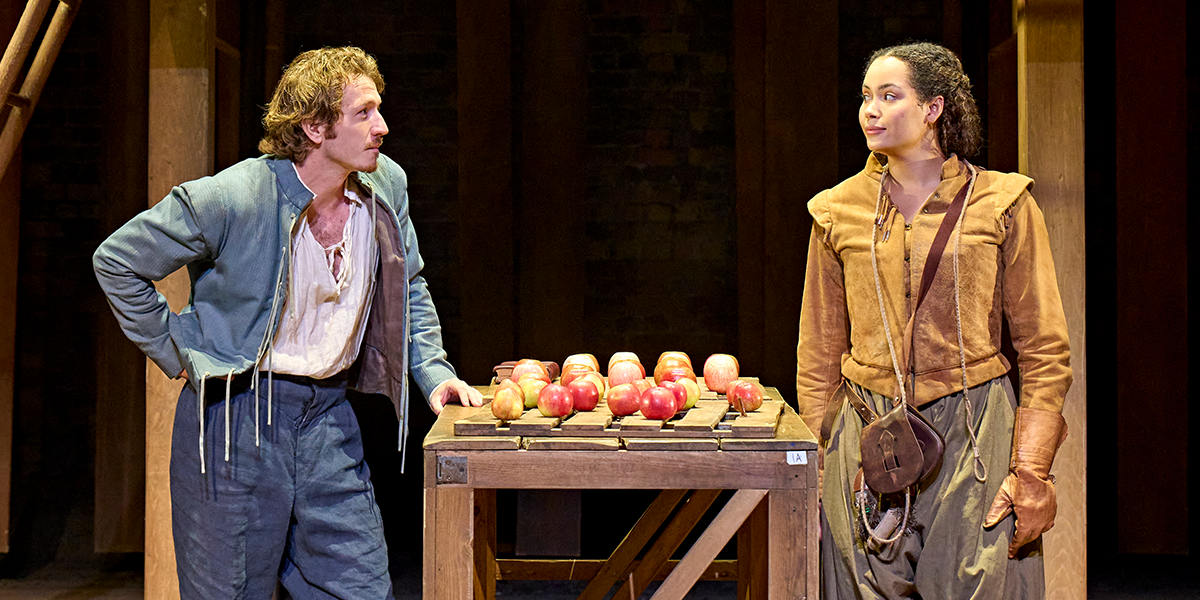 Tom Varey (William) and Madeleine Mantock (Agnes) face each other over a table covered in apples. Tom wears a blue jacket and breeches, Madeleine wears a brown jacket and skirt pulled into breeches. Photo by Manuel Harlan © RSC