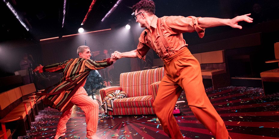 Two dancers holding hands as they move in a room that has sofas and chairs in.