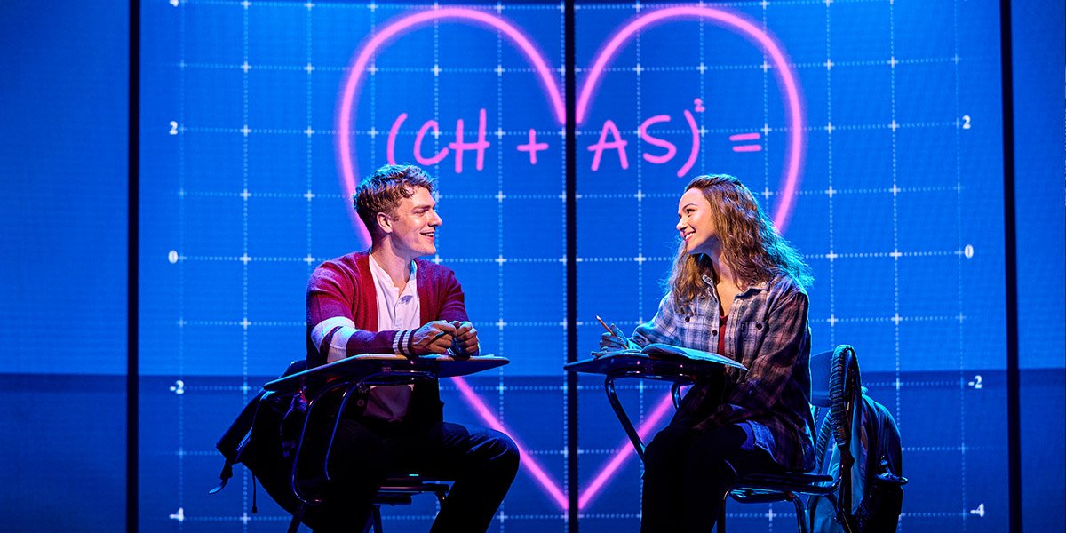 Daniel Bravo (Aaron) and Charlie Burn (Cady) sat at school desks opposite each other, in the background is a projection of a heart with their initials in.