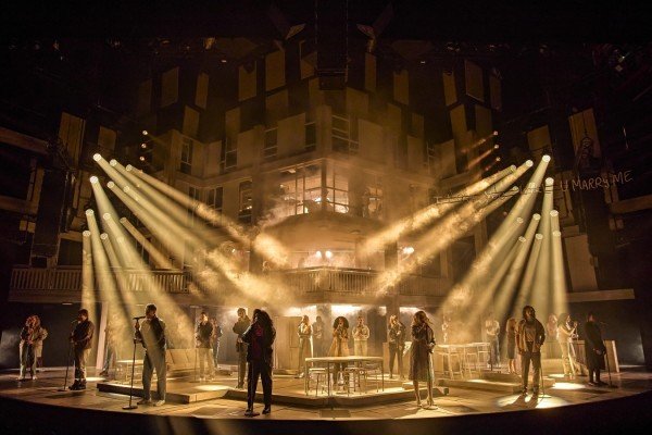 The cast of Standing at the Sky's Edge in the West End, lit in brown lighting. They stand across the stage, singing into microphones on stands.