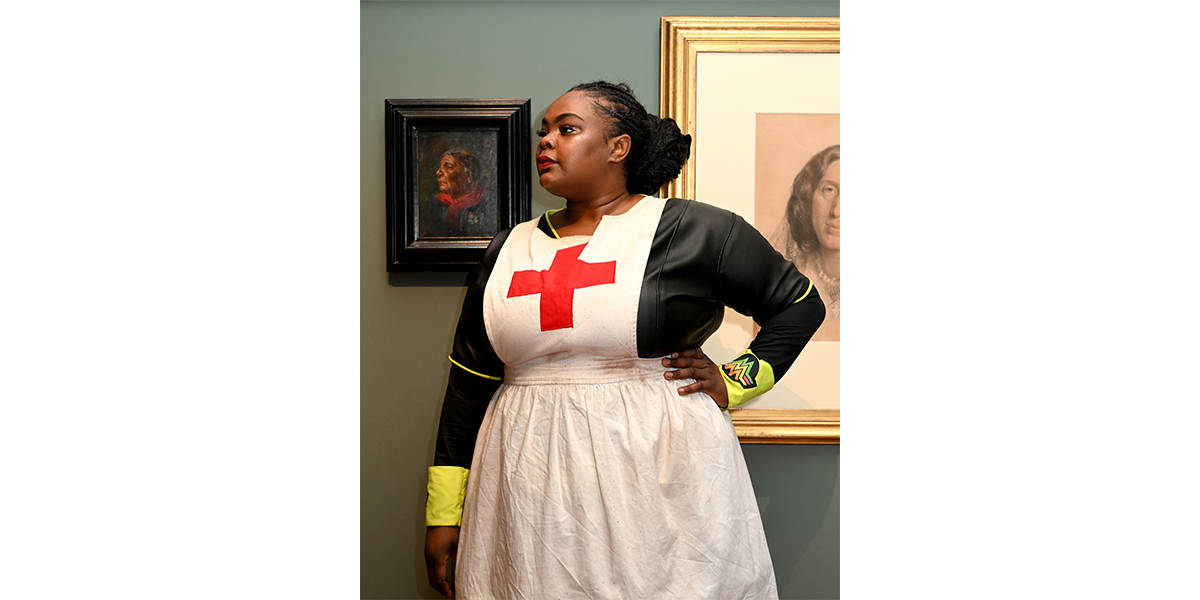 A woman stands wearing a white apron with a red cross on it, looking at a portrait