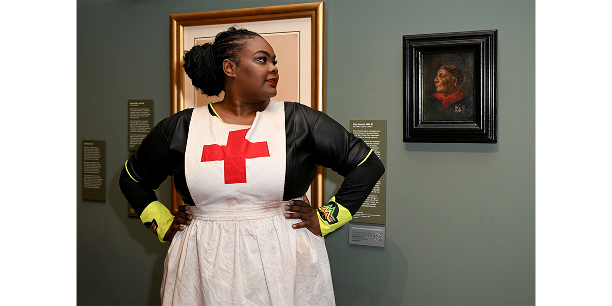 A woman stands wearing a white apron with a red cross on it, looking at a portrait