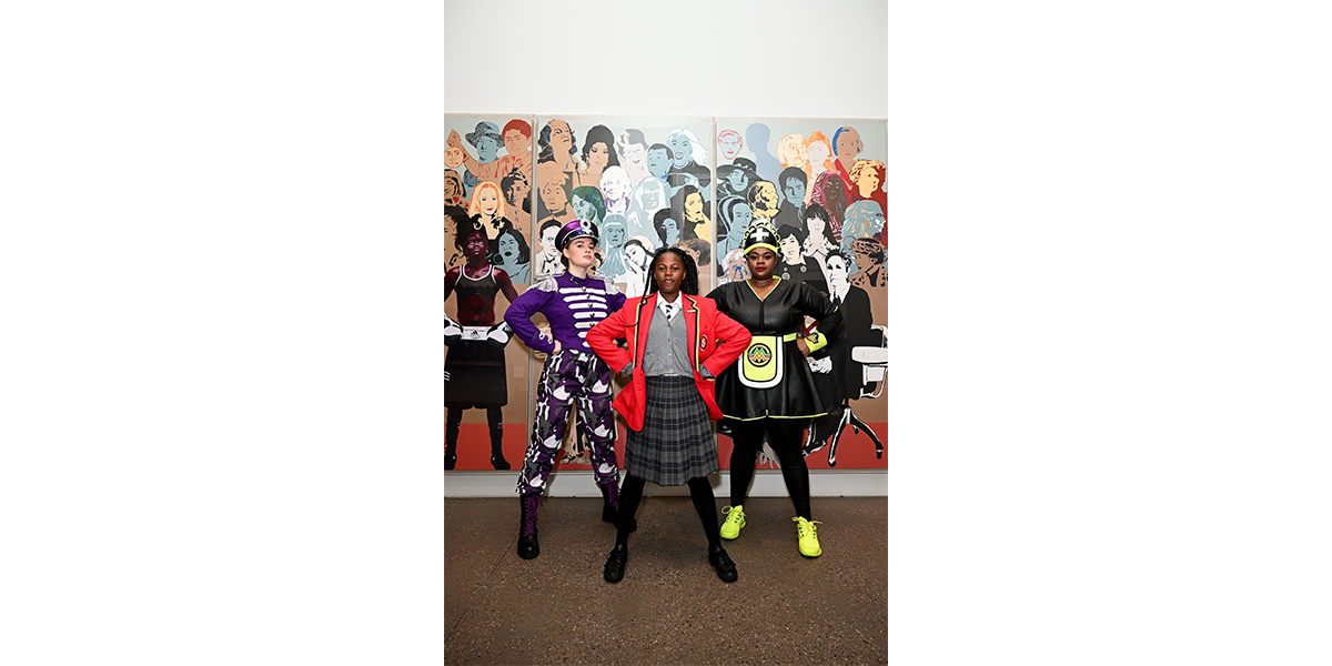 A woman wearing a black and neon green dress, a girl in a school uniform and a woman in a purple military outfit stand in front of a mural of hundreds of women's faces