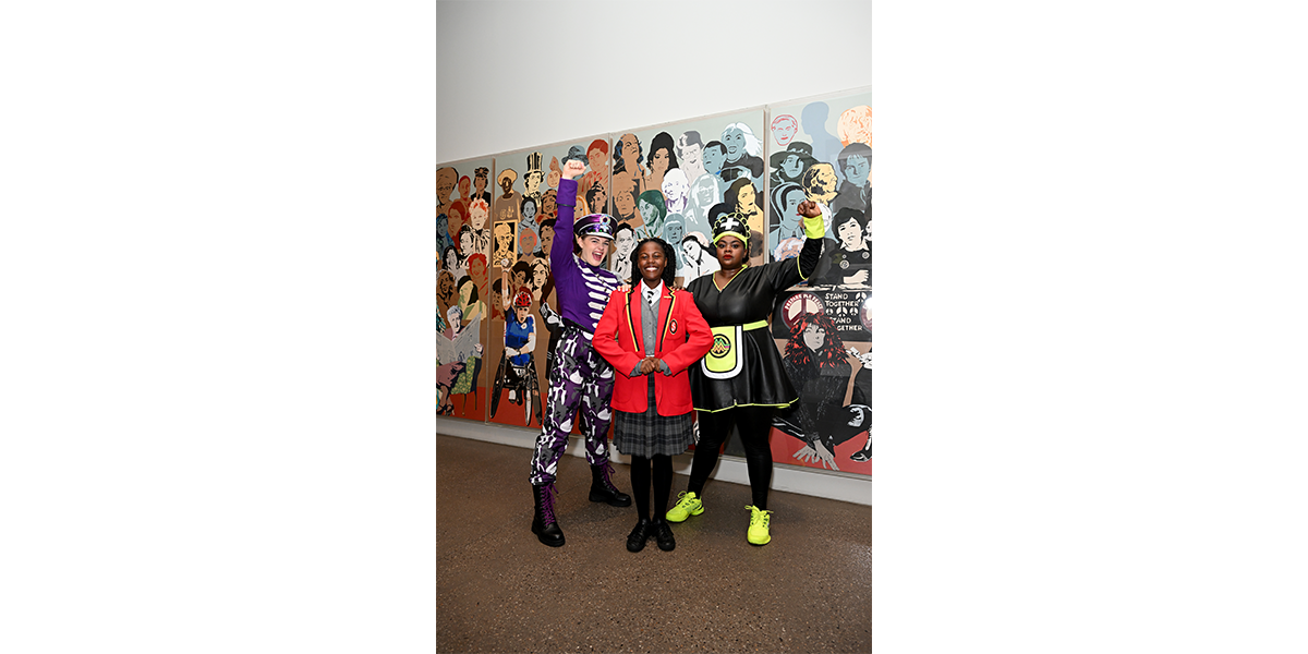 A woman wearing a black and neon green dress, a girl in a school uniform and a woman in a purple military outfit stand in front of a mural of hundreds of women's faces