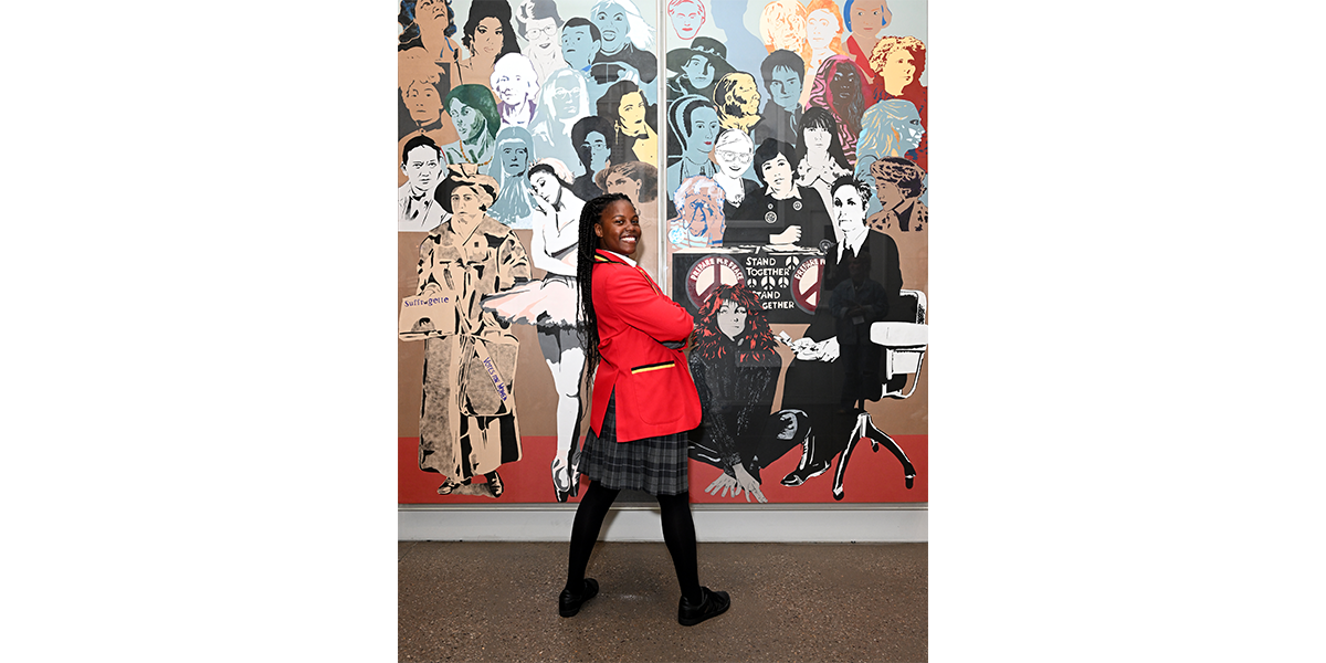 A girl in a school uniform stands in front of a mural of hundreds of women's faces