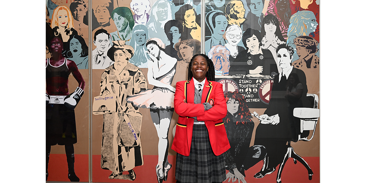 A girl in a school uniform stands in front of a mural of hundreds of women's faces