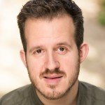 A close-up portrait of a man with short brown hair and a beard. He has a friendly expression and is wearing a green top, with a softly blurred outdoor background.