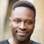 A close-up portrait of a man with short hair and a warm smile. He is wearing a black top, with a softly blurred background in an outdoor setting.