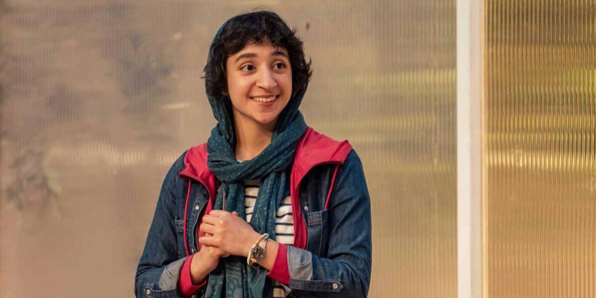 A young woman smiling warmly, wearing a headscarf, denim jacket, and a pink hooded jacket, with her hands clasped together.