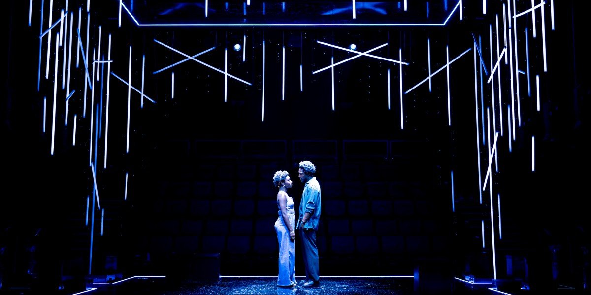 Two actors stand face to face on a black stage lit with white tube lights, suspended above their heads.