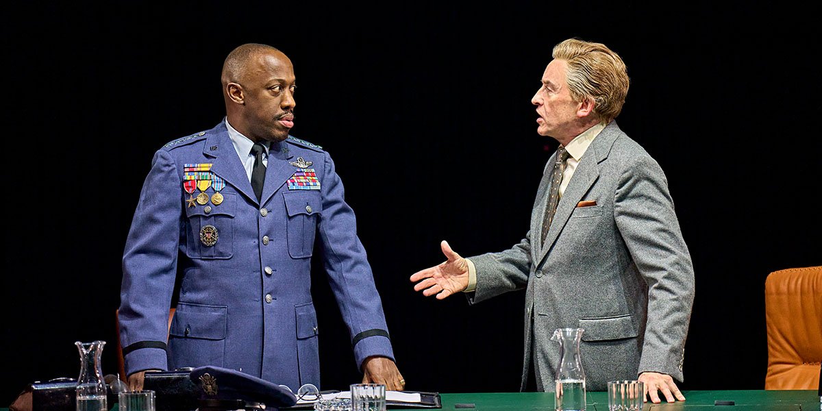 Giles Terera (General Turgidson) and Steve Coogan (President Muffley). On the right, Steve wears a grey suit and is talking to Giles. On the left, Giles wears a decorated navy army suit and is looking at Steve with concern.