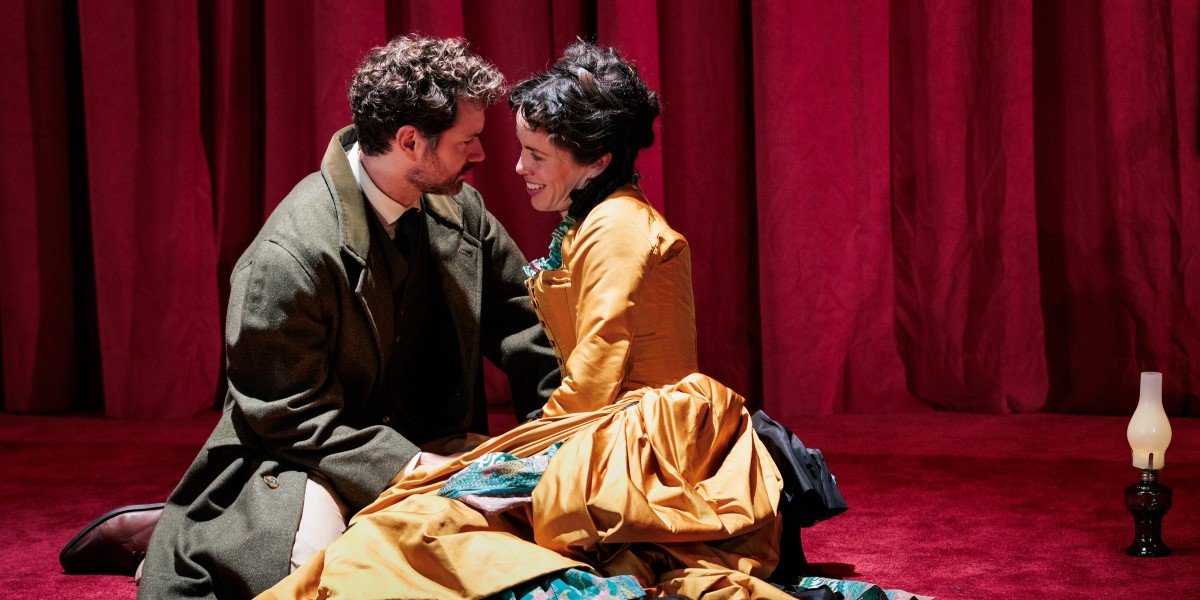 Two actors in Victorian dress sit on the floor, facing each other in a loving embrace